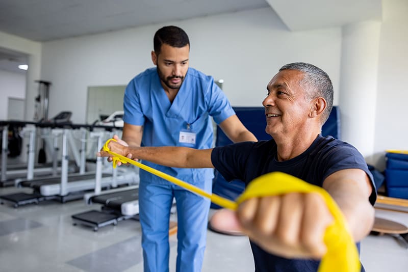 Mature Latin American man doing physical therapy exercises using a stretch band with the assistance of his therapist